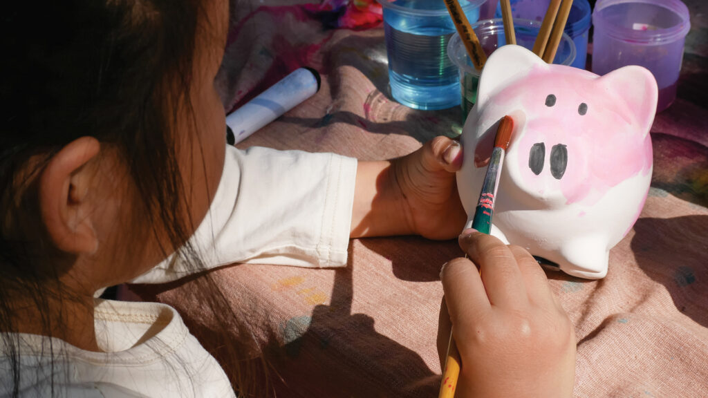 A child paints a white piggy bank pink using a brush at a table with art supplies.