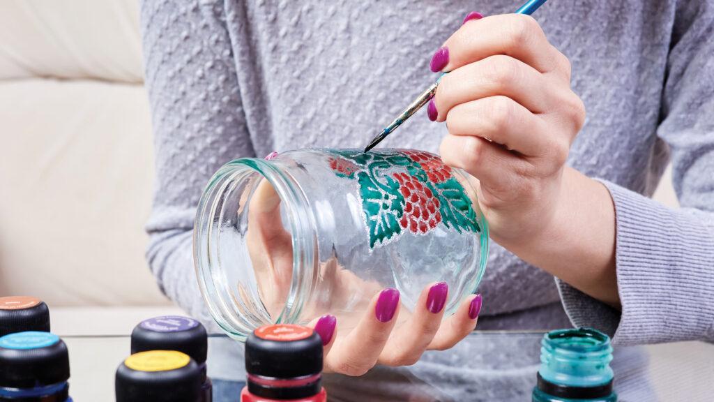 Person painting colorful flowers on a glass jar with a brush, surrounded by open paint containers.