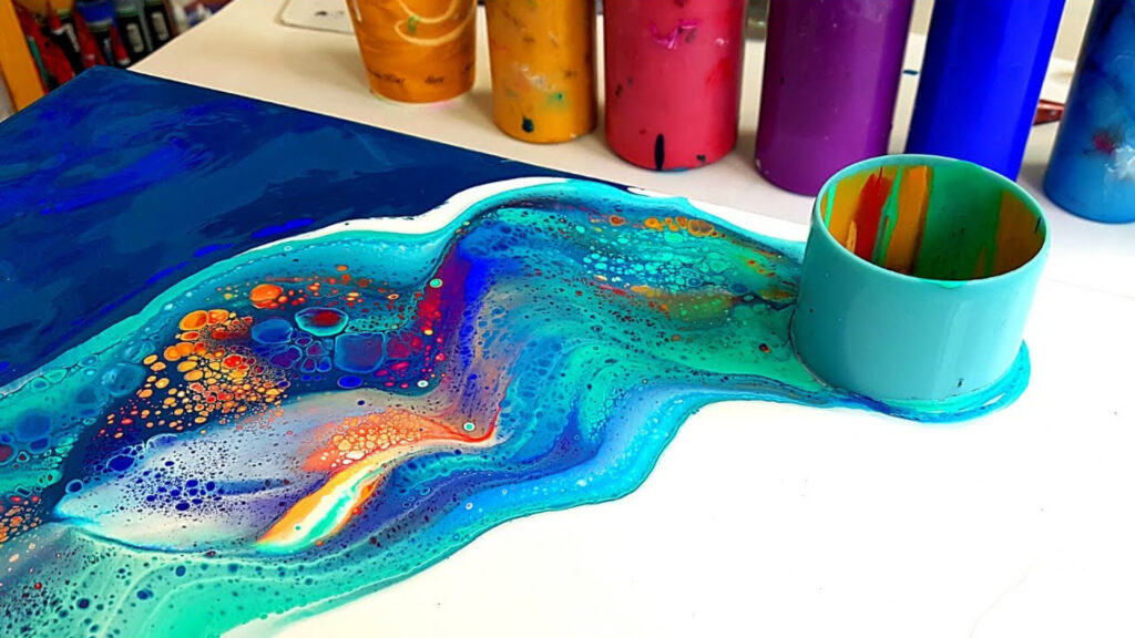 Abstract artwork in vibrant blues, turquoise, and orange with a cup and paint bottles in the background.
