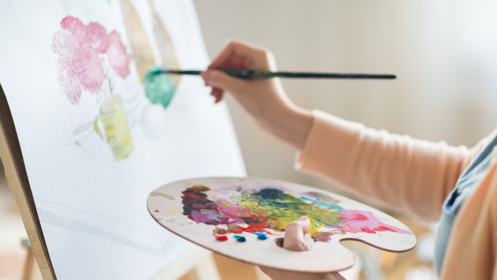 A close-up of a person painting on a canvas, holding a palette full of vibrant colors, with a brush in hand.