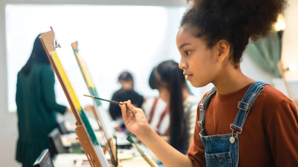 A young girl in overalls paints on a canvas in an art classroom with other students in the background.
