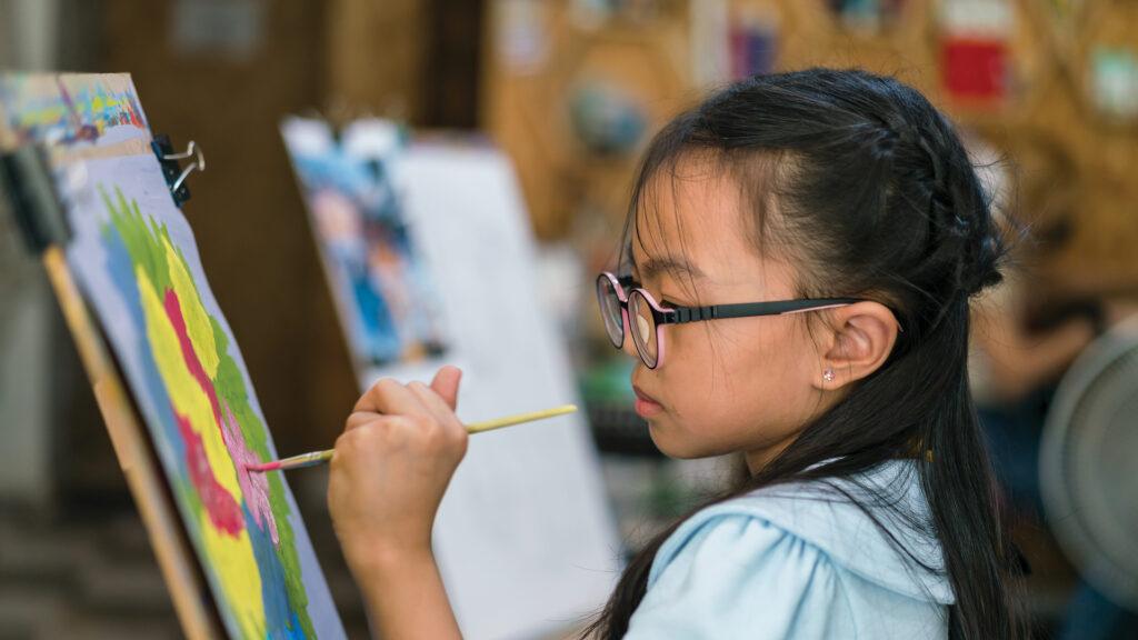 A young girl with glasses paints on a canvas at an easel, concentrating on her colorful artwork.