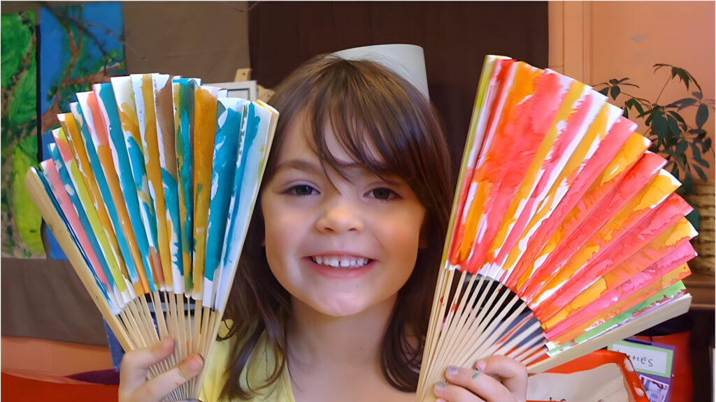 Smiling child holding two colorful, hand-painted paper fans, one in each hand, in a cheerful indoor setting.
