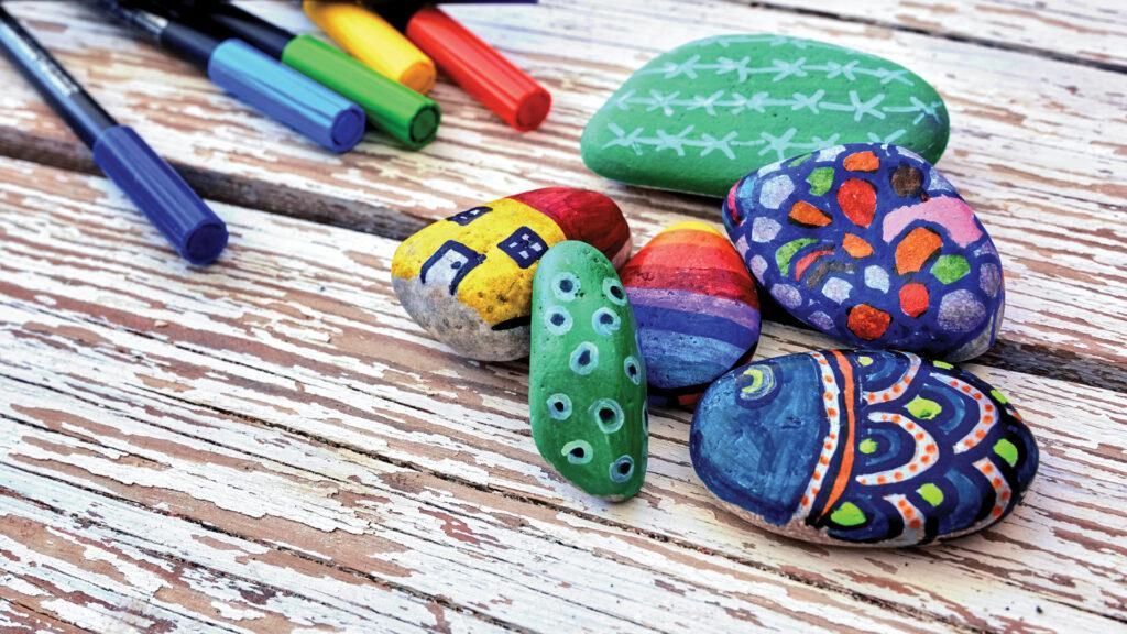 Colorful painted stones with various designs on a wooden surface, surrounded by markers.
