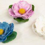 Three ceramic flowers in blue, pink, and white with yellow centers and green leaves on a light surface.
