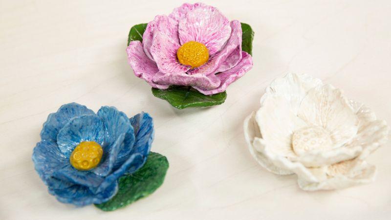 Three ceramic flowers in blue, pink, and white with yellow centers and green leaves on a light surface.