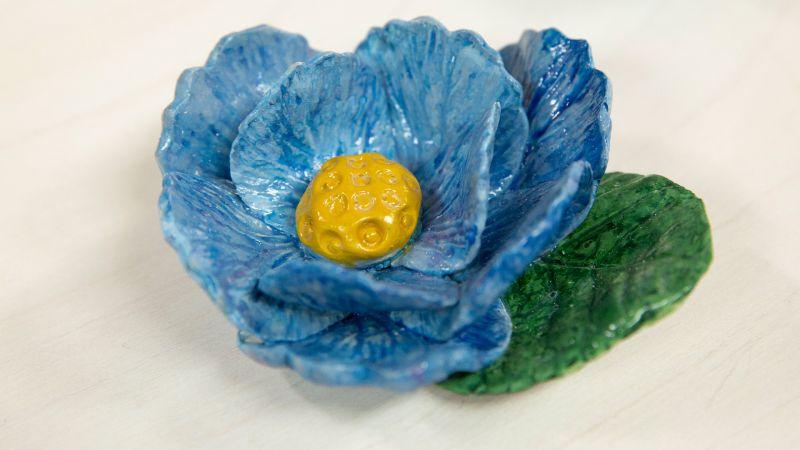 A blue ceramic flower with a yellow center and a green leaf on a light surface.