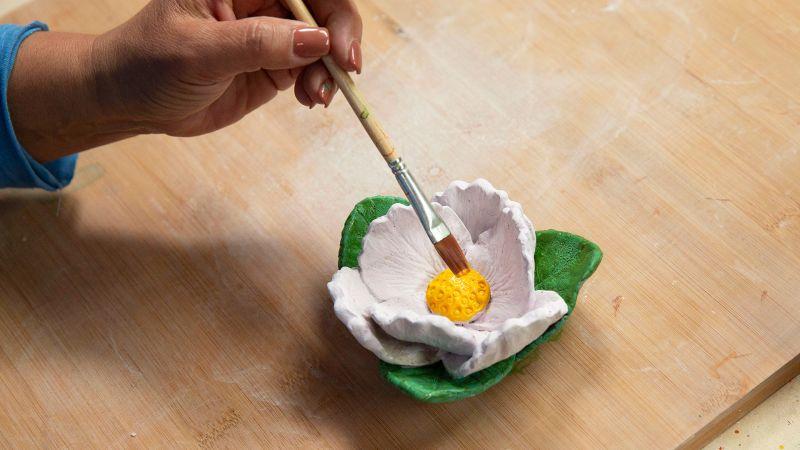 A hand painting the yellow center of a white clay flower with green leaves on a wooden surface.