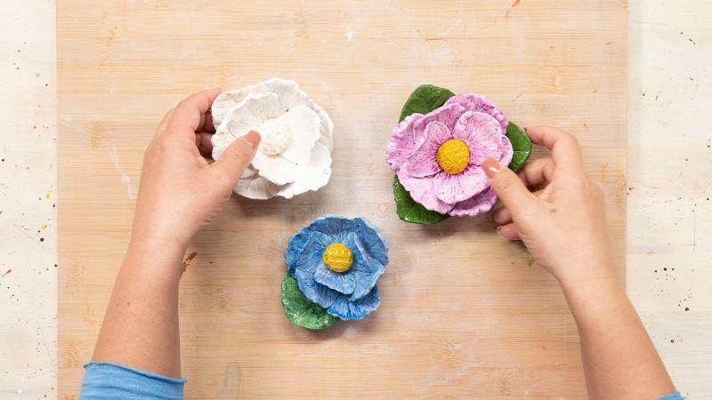 Hands arranging three colorful clay flowers on a light wooden surface, one white, one blue, and one pink.