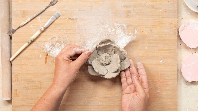 Hands shaping a clay flower on a wooden surface, with brushes and tools nearby.