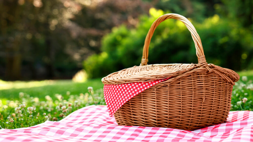 Wicker picnic basket with a red checkered cloth on a matching blanket, set on grass outdoors.