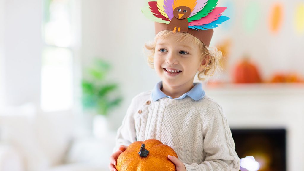 Child wearing a colorful feathered paper hat and holding a small pumpkin, smiling in a festive, bright room.