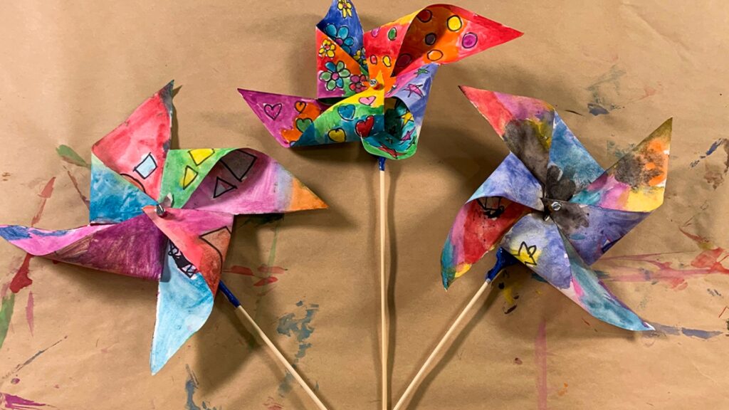 Three colorful, hand-painted paper pinwheels on sticks, arranged on a brown, paint-splattered background.