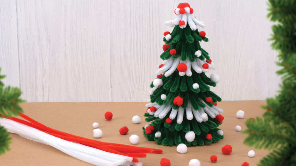A mini Christmas tree made of pipe cleaners and pom-poms sits on a table, with craft supplies nearby.