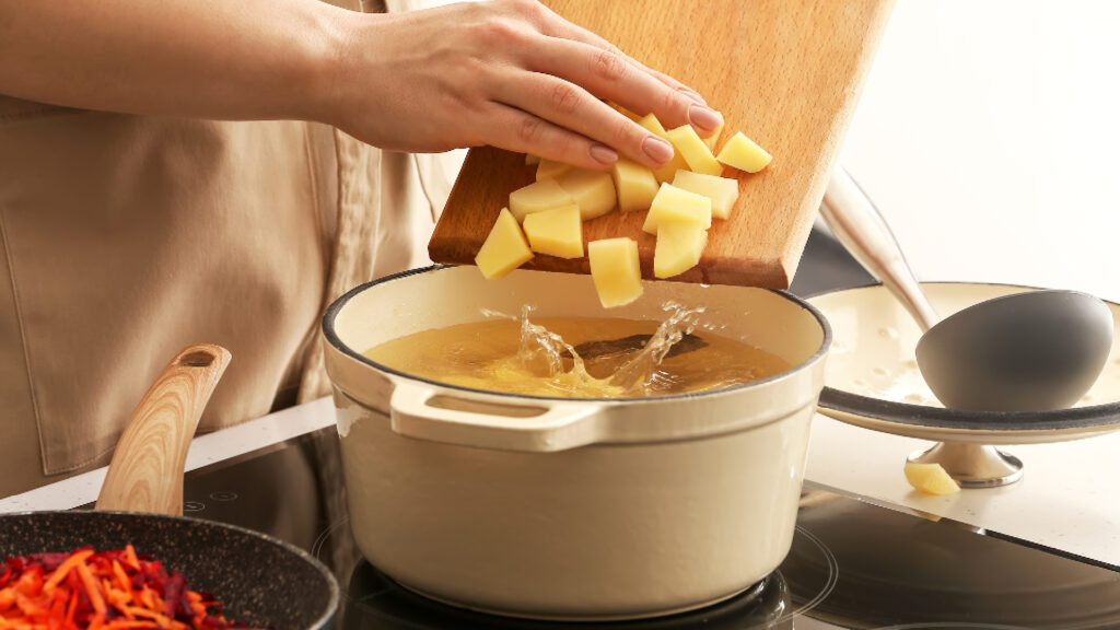A person adds diced potatoes from a cutting board into a pot of boiling water on a stove.