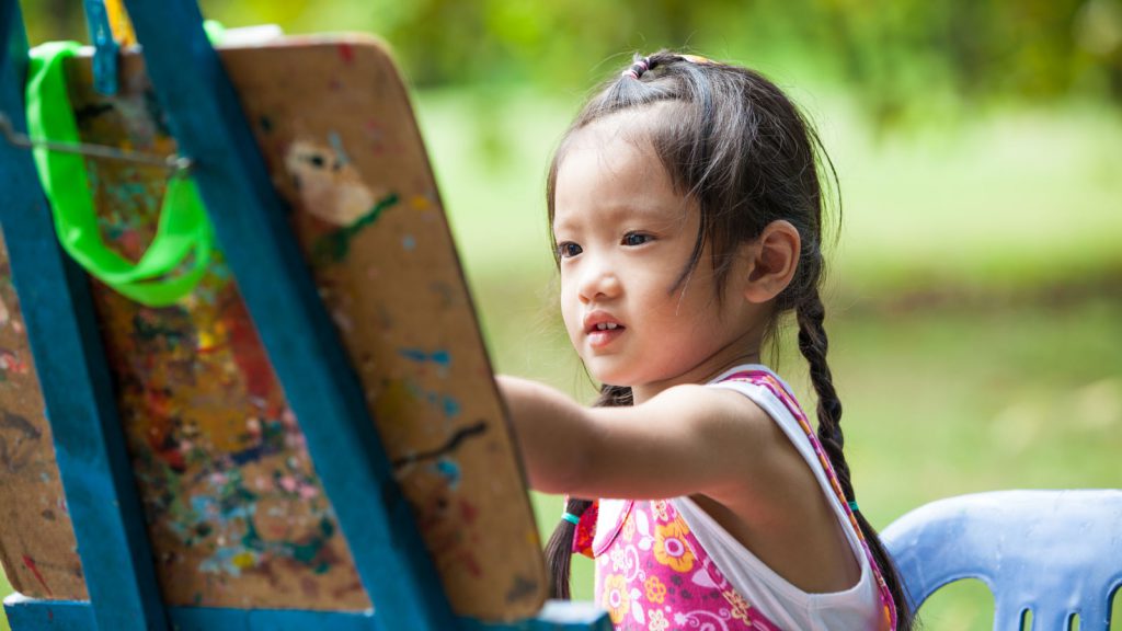 Young girl with pigtails painting on an easel outdoors, wearing a pink floral dress and sitting on a blue chair.