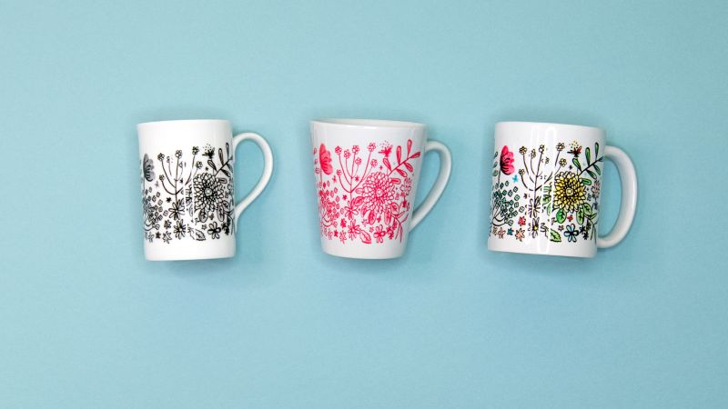 Three white mugs with floral designs in black, pink, and multicolor are placed against a light blue background.