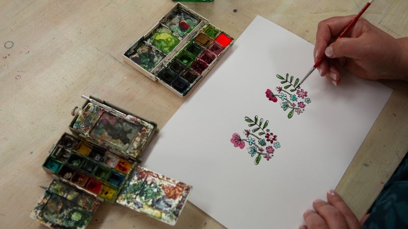 A person painting floral designs on white paper with watercolor paints and brushes on a table.