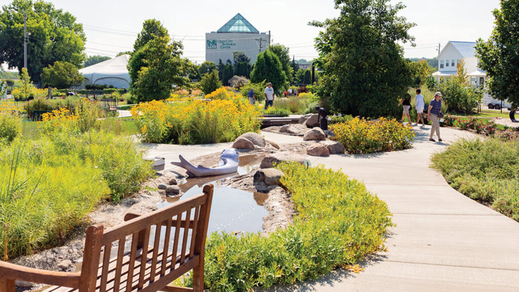 A sunny botanical garden with flowers, a bench, a small stream, walking paths, and visitors exploring the area.