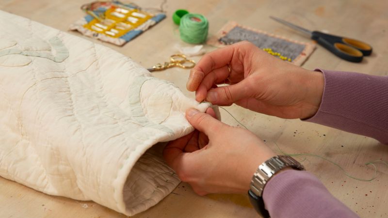Close-up of hands stitching a white quilt with green thread on a table, with sewing tools nearby.