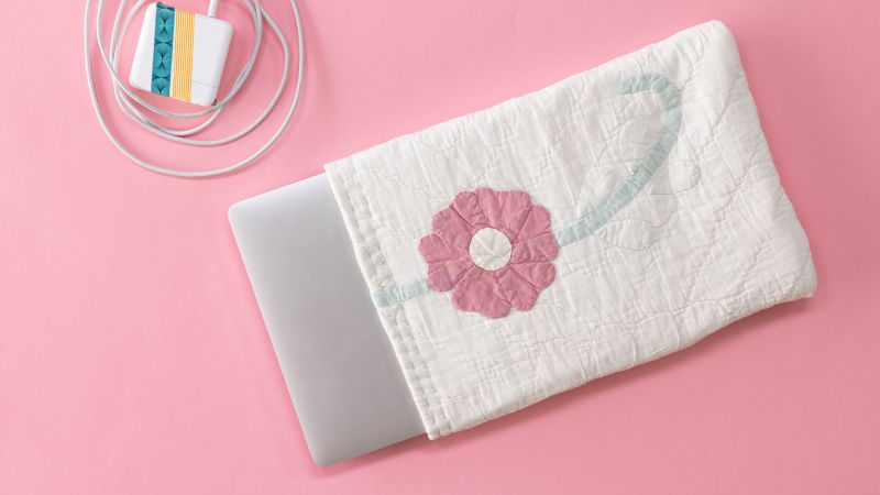Laptop partially in a quilted floral sleeve on a pink surface, with a colorful charger beside it.