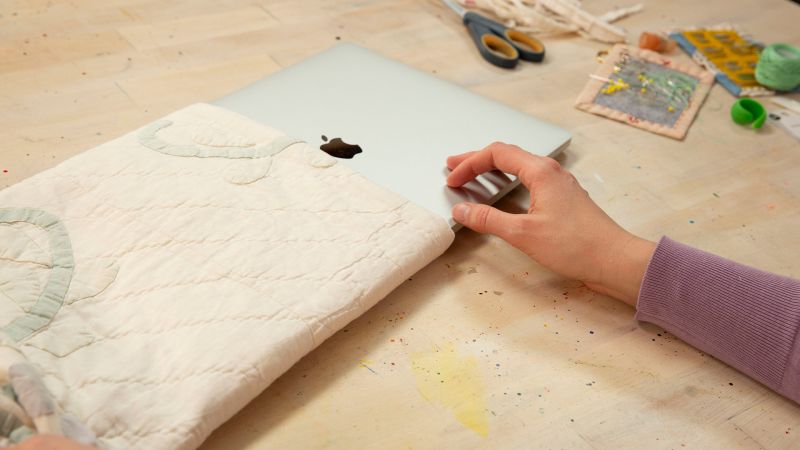 A person slides a laptop into a quilted sleeve on a craft table with scissors, pins, and various sewing materials.