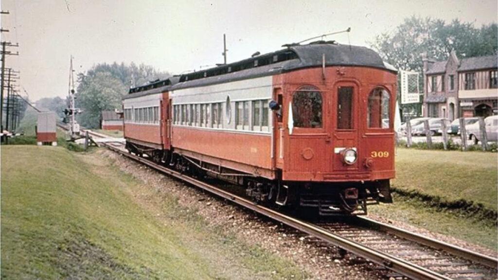 A vintage red and cream electric train travels on tracks through a grassy area near buildings and parked cars.