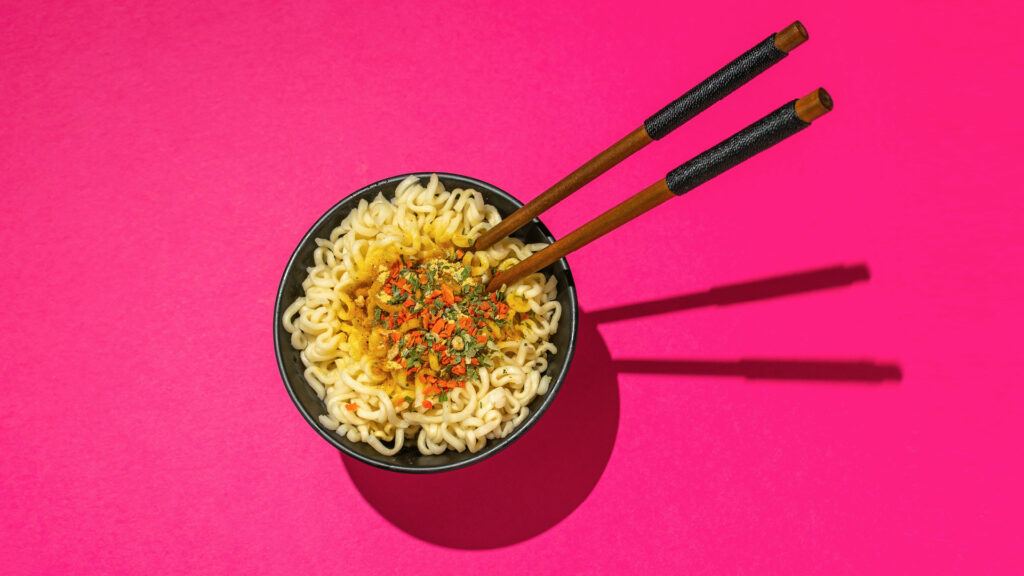 Bowl of ramen noodles with chopsticks on a bright pink background.