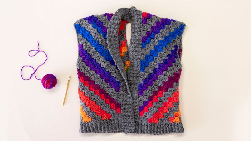 A colorful granny square crochet vest with a gray border, a crochet hook, and a ball of purple yarn.