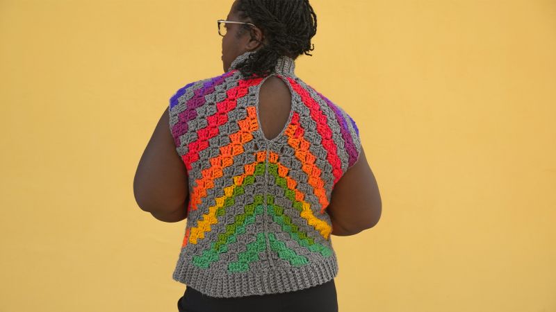 Person with braids wearing a colorful crocheted top stands against a yellow background, facing away from the camera.