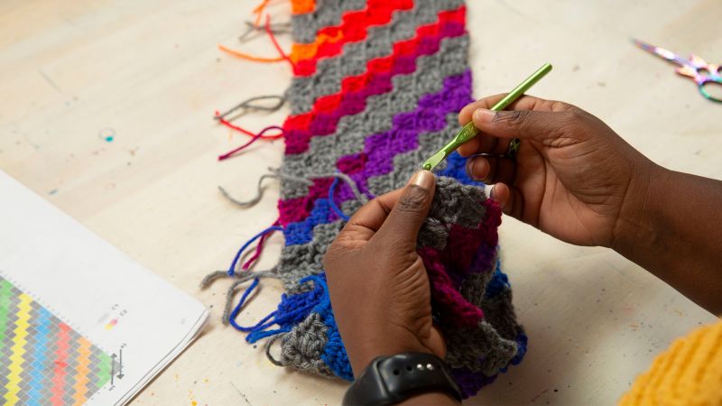 Hands crocheting a colorful pattern with a green hook, next to a crochet pattern chart and scissors on a table.