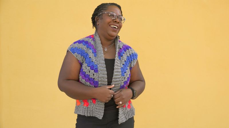 A smiling person wearing a colorful crocheted vest stands against a yellow background.