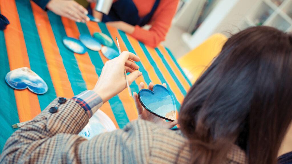 Person painting a blue heart-shaped object at a table covered with a colorful striped cloth.