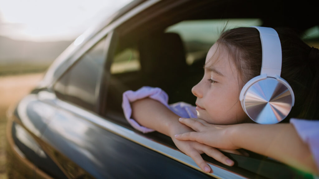 A girl wearing headphones looks out a car window, leaning her arms on the door and enjoying the scenery.