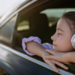 A girl wearing headphones looks out a car window, leaning her arms on the door and enjoying the scenery.