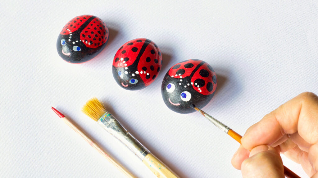 Three rocks painted as ladybugs, three paintbrushes, and a hand painting one rock on a white background.