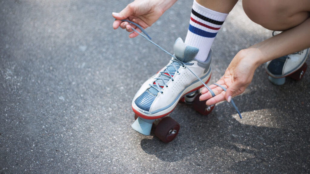 Person tying the shoelaces of a white roller skate on an asphalt surface, wearing striped socks.