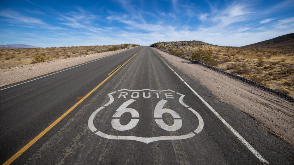 A deserted Route 66 highway stretches into the distance through a dry, desert landscape under a blue sky.