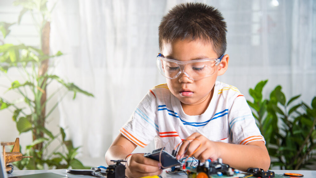 Young child wearing safety goggles and a striped shirt, focused on assembling a small electronic project indoors.