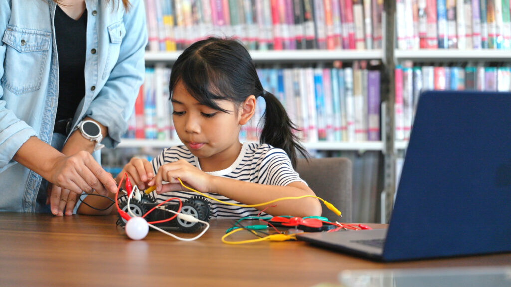 A young girl works on a STEM project with wires, next to an adult and a laptop in a library.