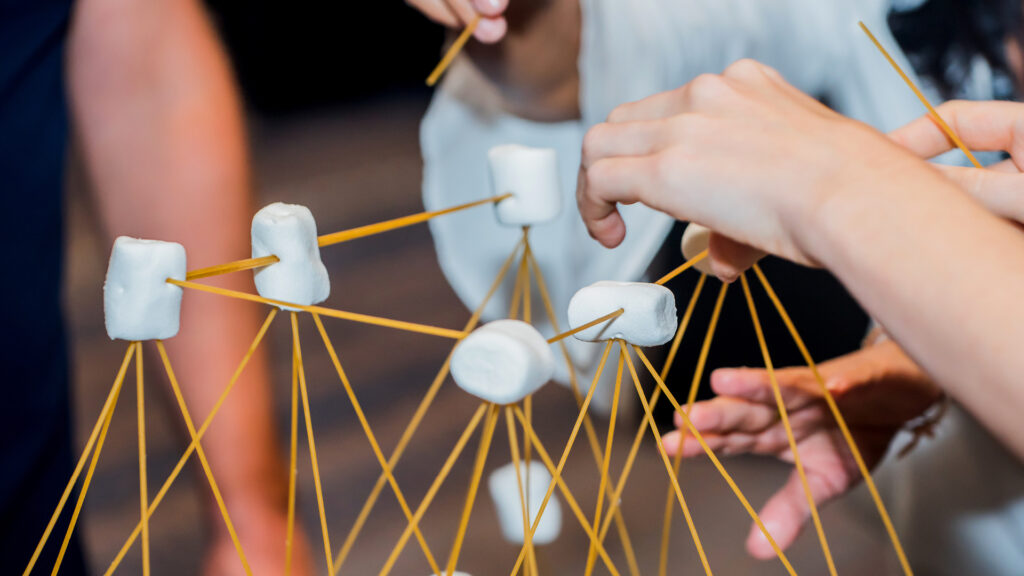 Hands building a structure with spaghetti sticks and marshmallows during a team activity.