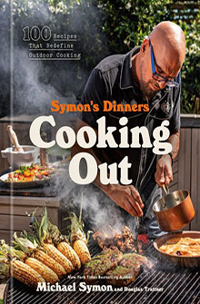 A man cooks outdoors by a grill with vegetables and corn; cover of "Symon's Dinners Cooking Out" cookbook.