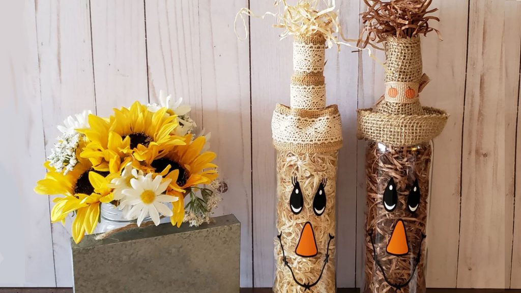 Two bottles decorated as scarecrows next to a vase of sunflowers and daisies on a wooden background.