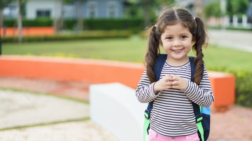 Smiling young girl with pigtails and a backpack standing outdoors in a park or schoolyard.