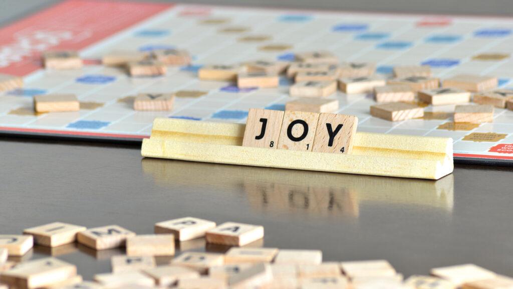Scrabble letter tiles spell "JOY" on a rack, with a game board and scattered tiles in the background.
