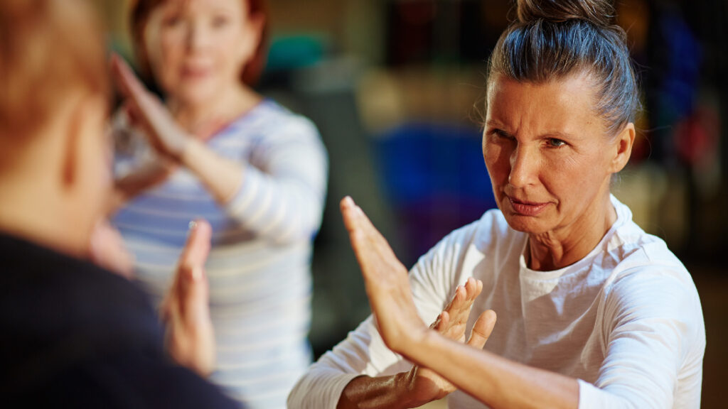 A focused woman practices martial arts with her hands extended, another person mirroring her movements in the background.