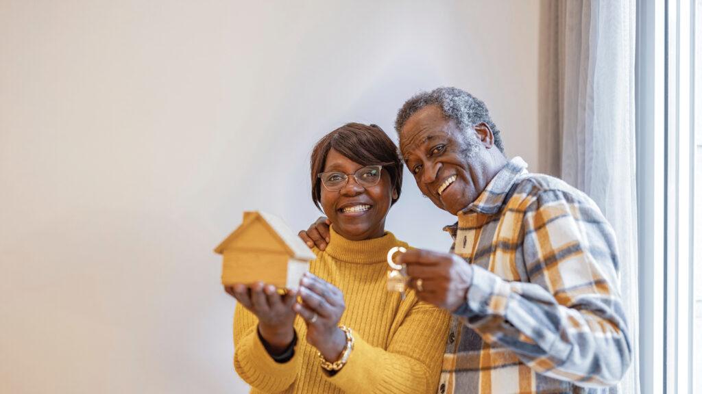 Smiling couple holding a small wooden house and keys, celebrating a new home by a bright window.