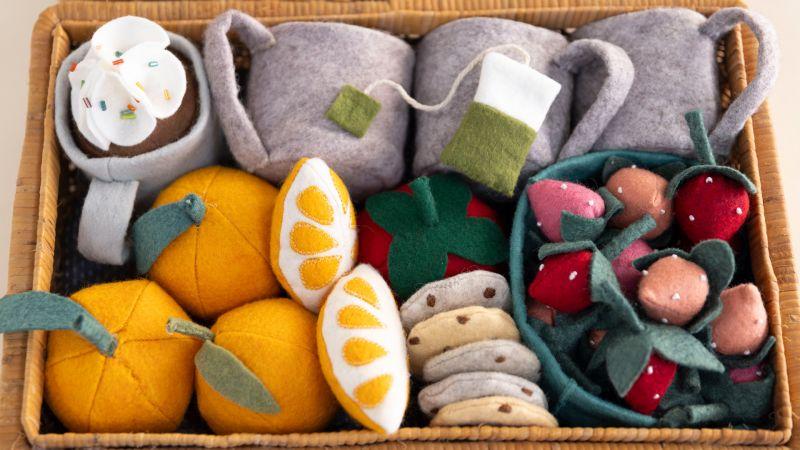 A basket filled with felt toy fruits, tea cups, cookies, and desserts in bright colors.