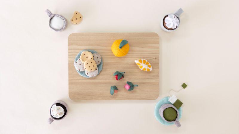 Felt cookies, fruit, and drinks on a light wooden board with cups and treats arranged around it.