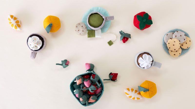 Colorful felt tea set with cups, cookies, fruit, and strawberries arranged on a light background.